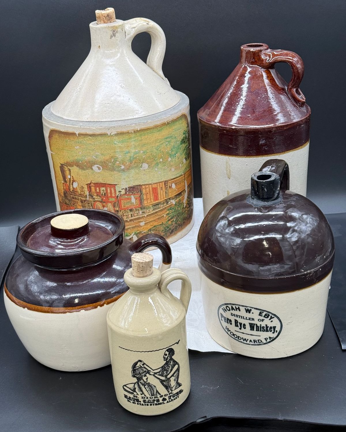 Five antique stoneware jugs arranged together, including a tall white jug with a transferred steam locomotive scene, a two-tone brown shoulder jug, a Noah W. Eby Pure Rye Whiskey jug from Woodward Pennsylvania, a round brown-lidded stoneware crock, and a small white figure-decal jug.