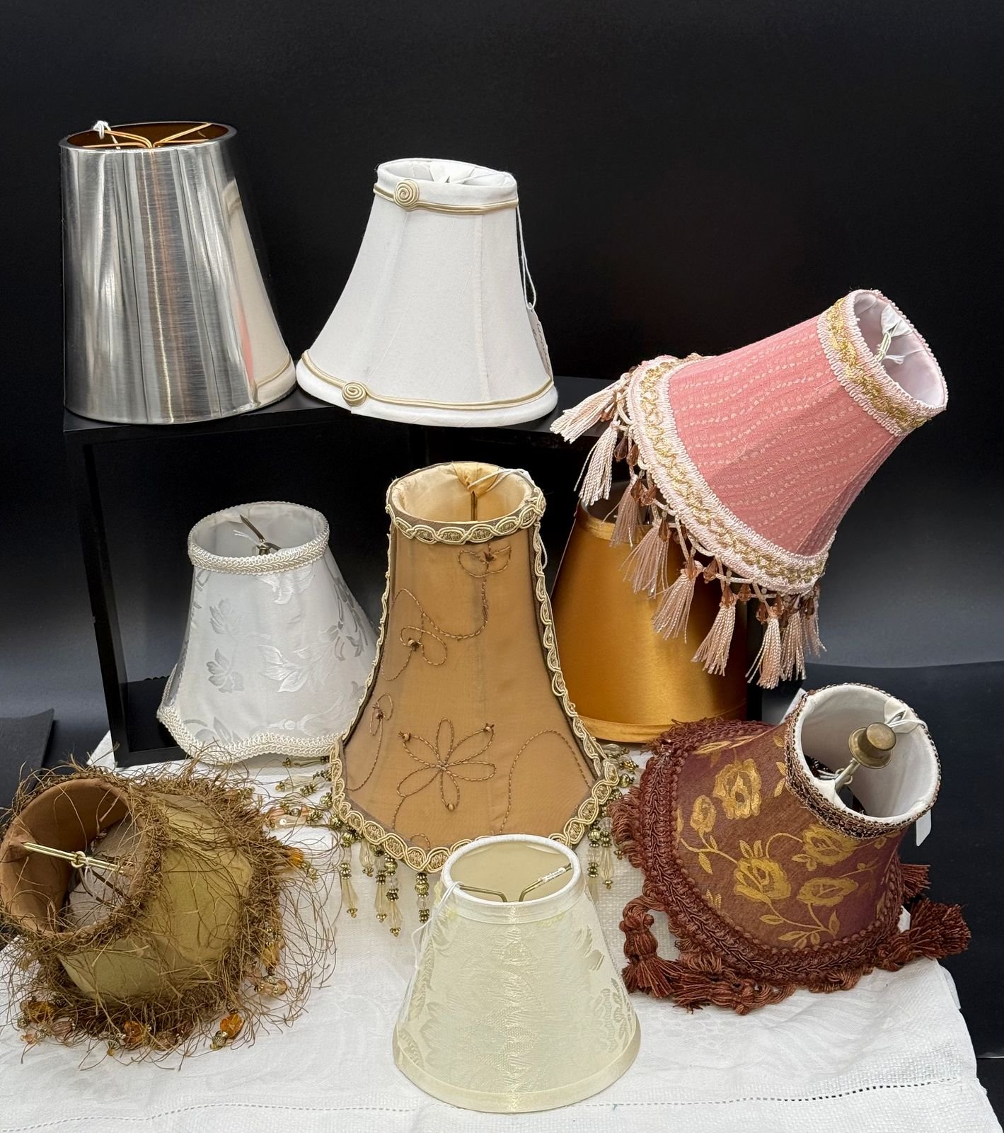 A grouping of nine vintage clip-on chandelier and sconce shades in varied fabrics and trims: silver satin, ivory silk, dusty rose with beaded fringe, gold embroidered, white damask, gold wirework, burgundy floral with bullion fringe, and cream embossed