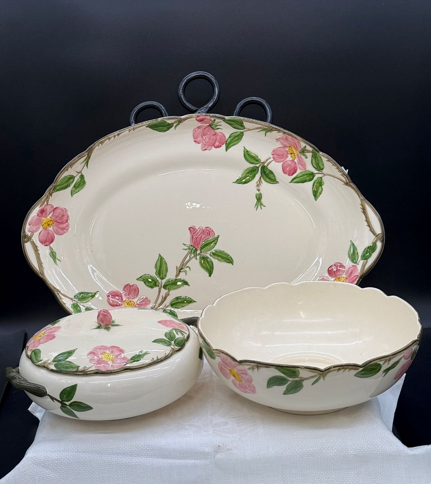 Three vintage Franciscan Desert Rose serving pieces: an oval platter with iron wall hanger, a lidded round bowl, and a scalloped serving bowl, all in cream with hand-painted pink roses and green leaves