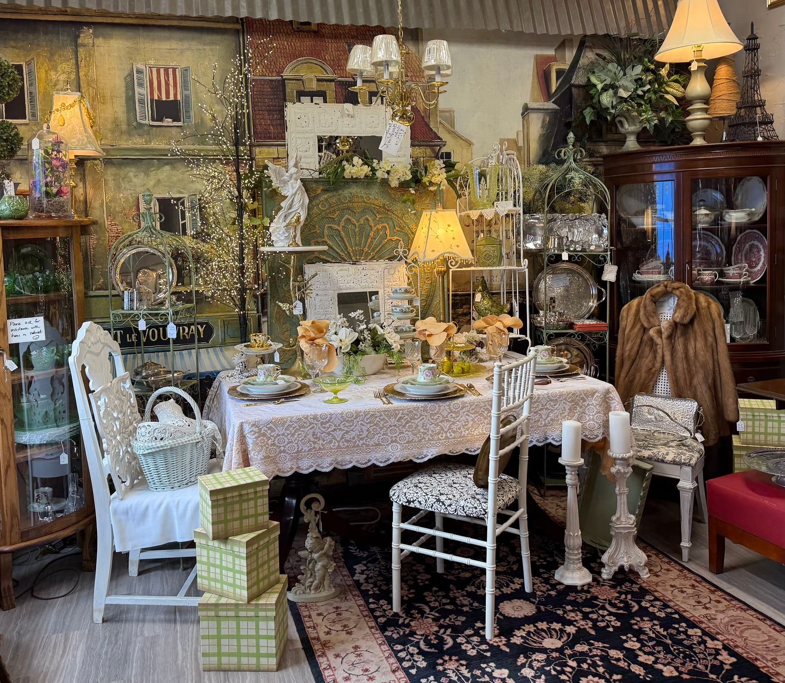 A French country–inspired table setting with ivory lace cloth, china place settings, tall candlesticks, and an apricot floral centerpiece, flanked by antique chairs in Pollyanna's Antiques shop.