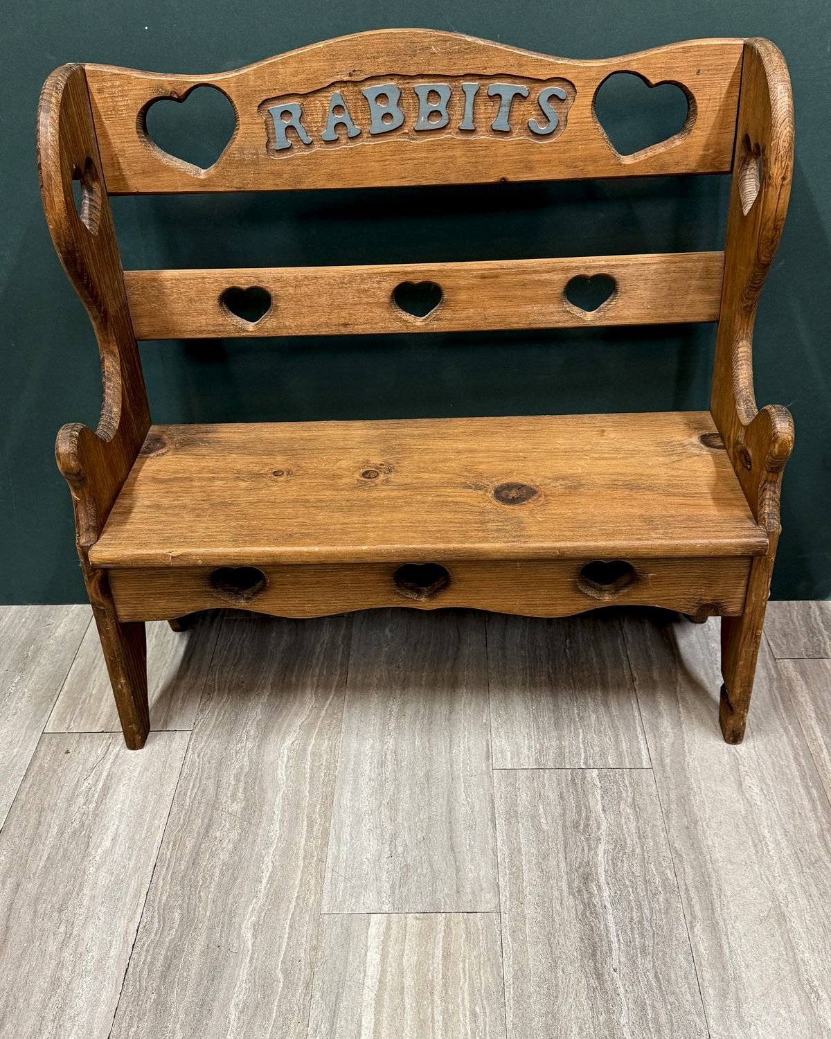 Small honey-toned wooden bench with scrolled arms, heart cutouts along the back rail and apron, and "RABBITS" carved across the top in blue lettering.
