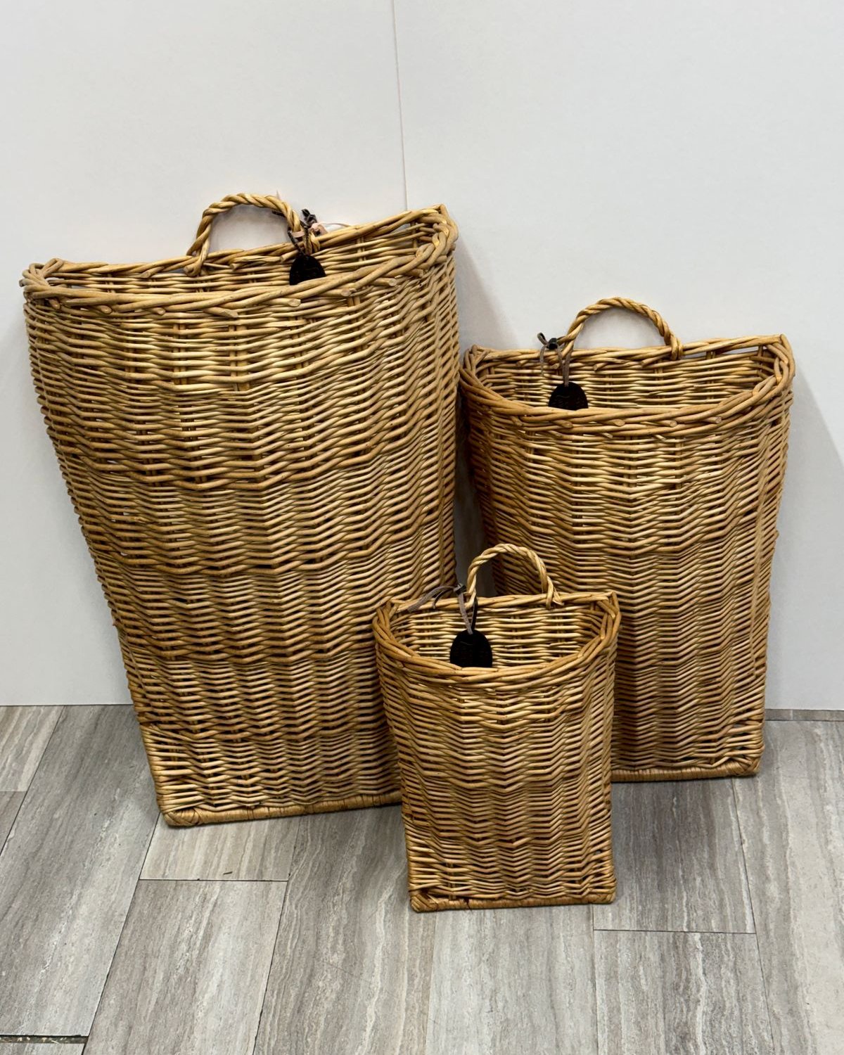 Three graduated woven wicker wall baskets with handles, hanging against a white wall above a wood floor.