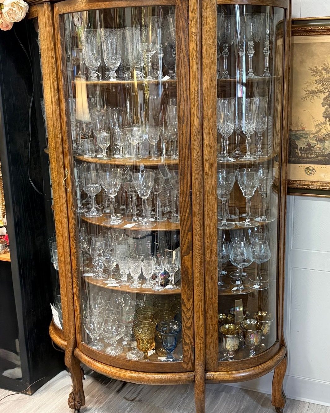 Antique tiger oak curio cabinet with curved bowed glass front, three wooden shelves filled with antique stemware and crystal, standing on carved lion paw feet.
