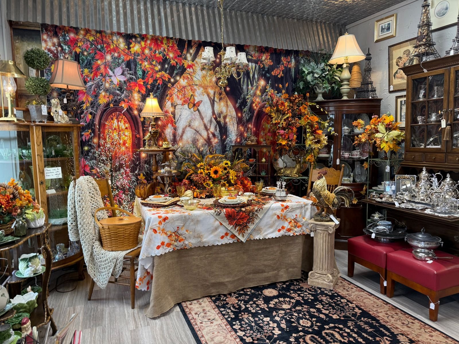 A fall table setting at Pollyanna's Antiques: a leaf-print table with autumn china, amber glass, and a sunflower centerpiece, framed by an autumn forest backdrop, lamps, and china cabinets.