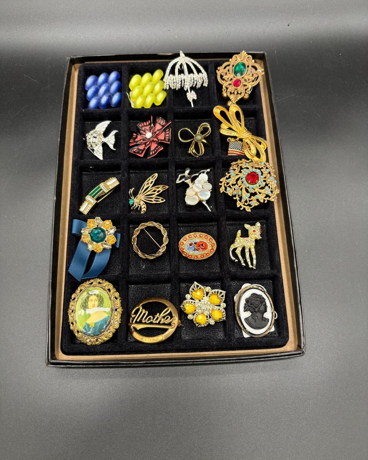 A black velvet jewelry tray of twenty compartments, every one filled with a vintage brooch, including a rhinestone umbrella with fringe, a silver angelfish, a red and black enamel pinwheel, a gold ribbon bow with a small American flag, a dragonfly, a mother-of-pearl flower spray, a micromosaic oval, a rhinestone fawn, a portrait brooch of a woman in a blue dress, a gold Mother brooch, and a black cameo on white.