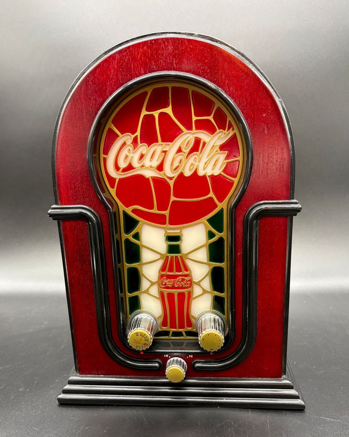 A 2002 Coca-Cola AM FM radio, twelve inches tall, built in the arched cathedral radio shape with a deep red cabinet, black deco trim and stepped base, three chrome and gold knobs, and a stained glass style front panel showing the Coca-Cola script above a contour bottle in red, green, and cream.