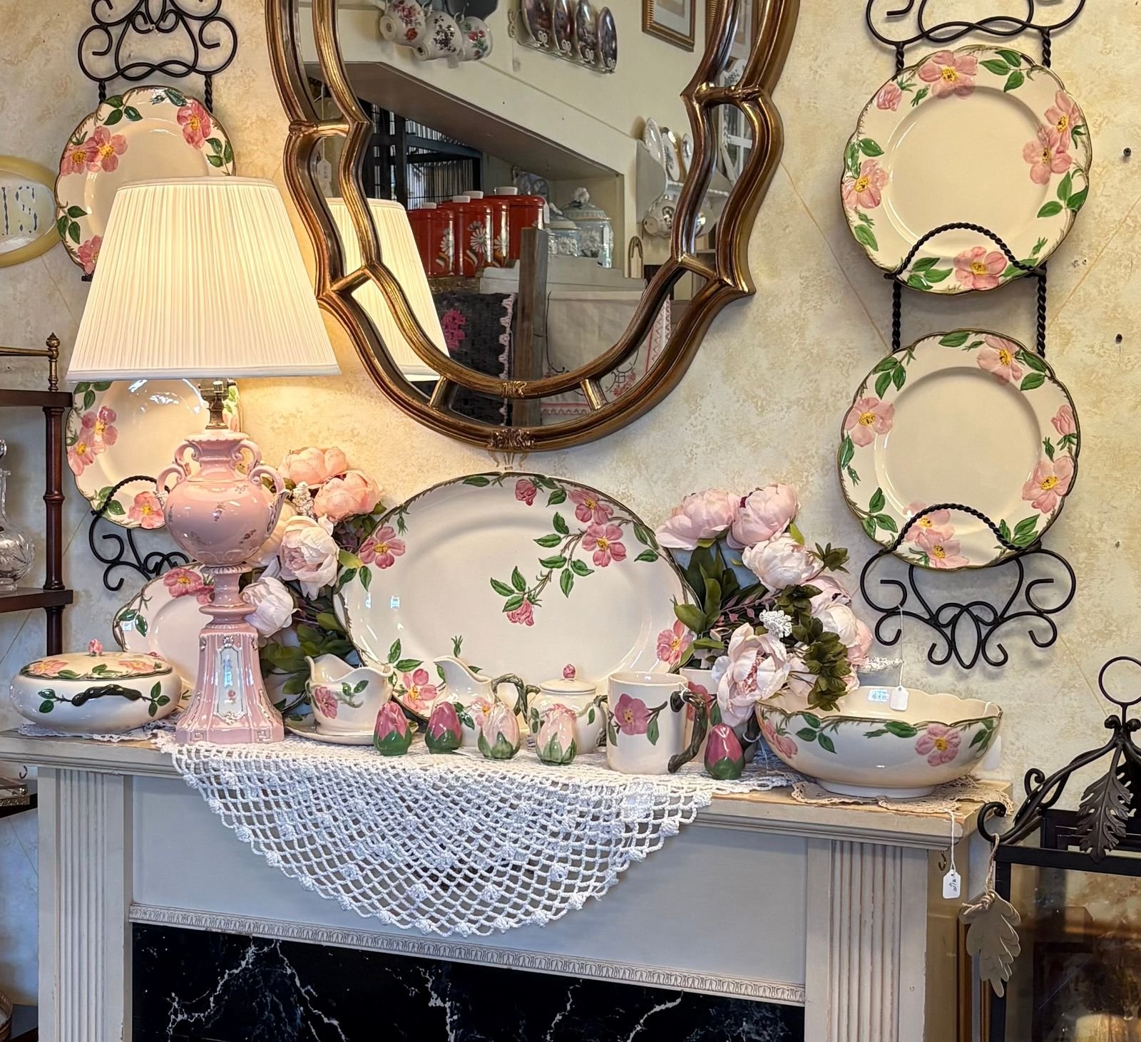 A mantle display of vintage Franciscan Desert Rose dinnerware, with dinner plates held on wrought iron wall racks, a large oval platter propped at center, and serving pieces along the shelf including a covered casserole, gravy boat, creamer, lidded sugar bowl, mug, a row of rosebud pieces, and a scalloped salad bowl, beneath a gold scalloped mirror with a crocheted lace runner spilling over the mantle edge.