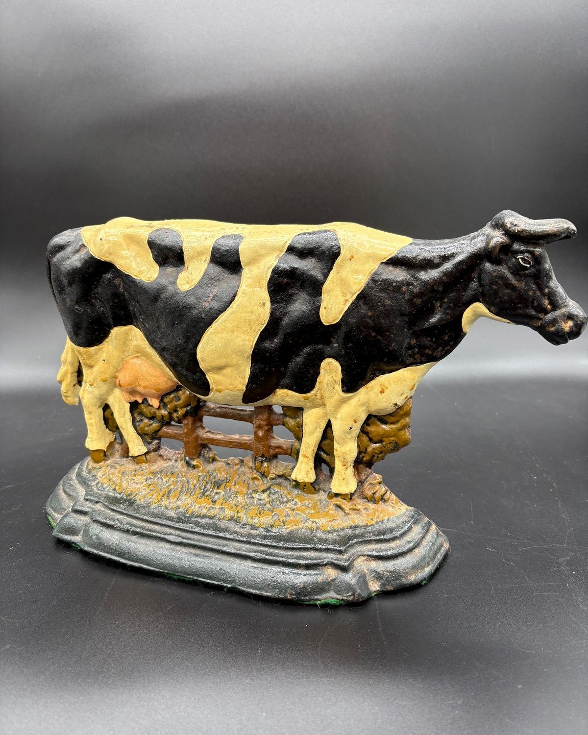 A hand-painted cast iron cow doorstop, black with soft cream markings, standing in profile at a brown cast rail fence with tufts of grass and shrubbery, mounted on a heavy oval base with a stepped molded edge, photographed against a gray backdrop.