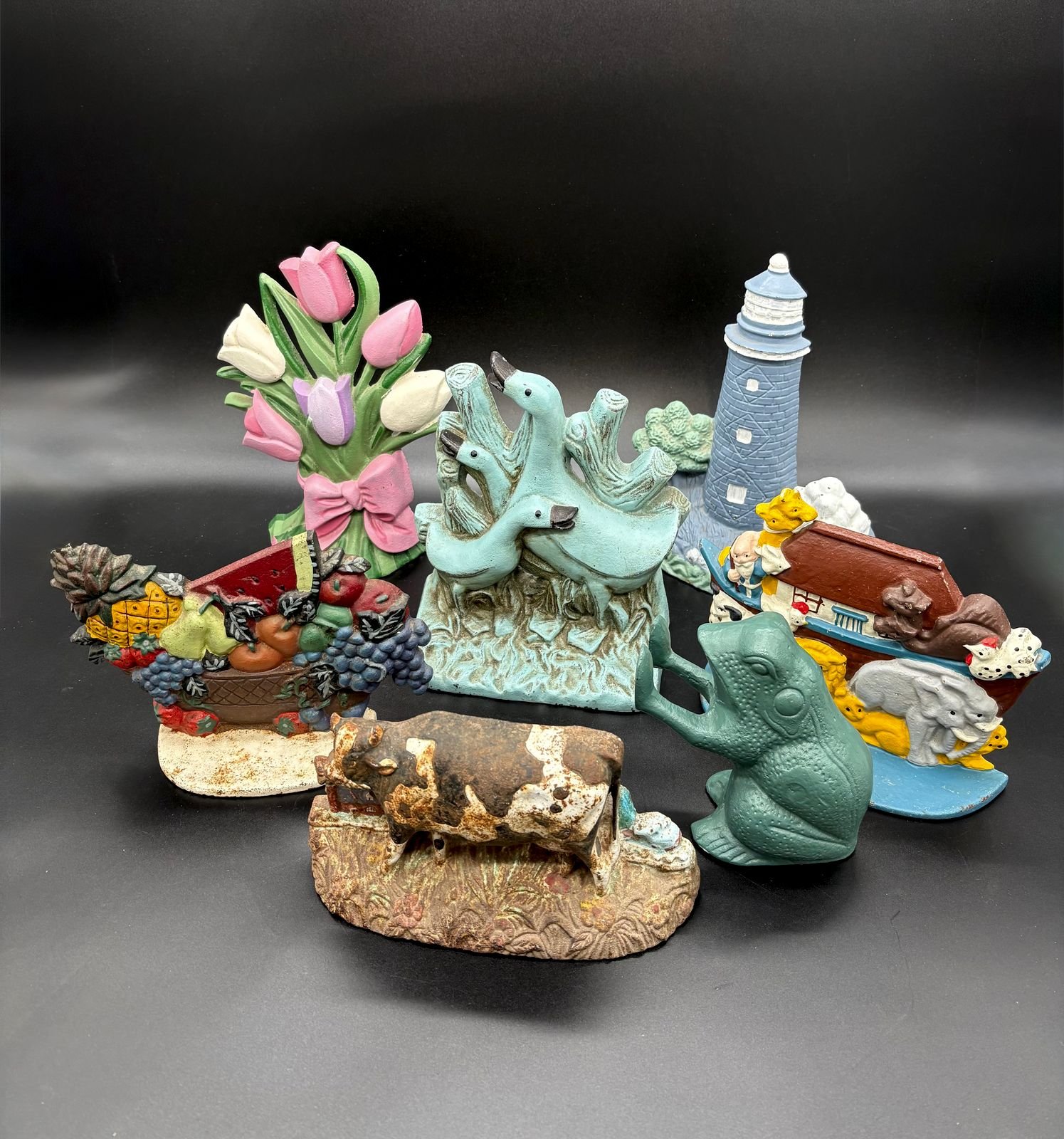 Seven vintage cast iron figural doorstops grouped on a gray backdrop: a pink and white tulip bouquet with a ribbon bow, three aqua geese at a tree stump, a blue and white lighthouse, a hand-painted cow at a fence, a green seated frog, a basket of painted fruit, and a Noah's Ark loaded with animals.