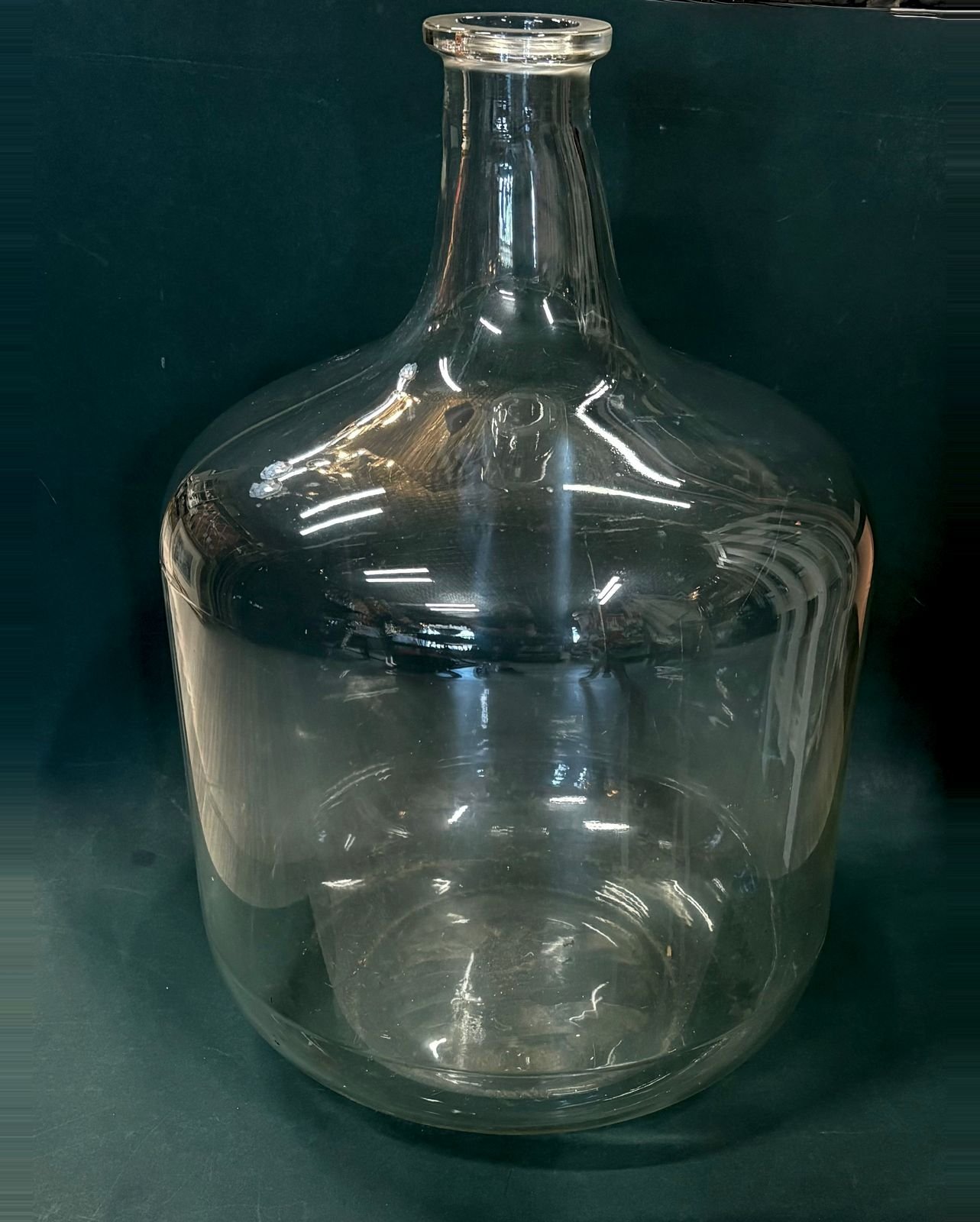 A large clear vintage Corning Pyrex glass carboy with a broad rounded body, tapered neck, and thick rolled lip, standing against a dark teal backdrop.