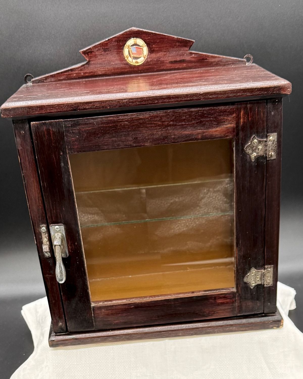 A 1920s dark stained wood wall-mount medicine cabinet with a peaked crown, a small round American flag badge, a glass door, and two glass shelves inside.