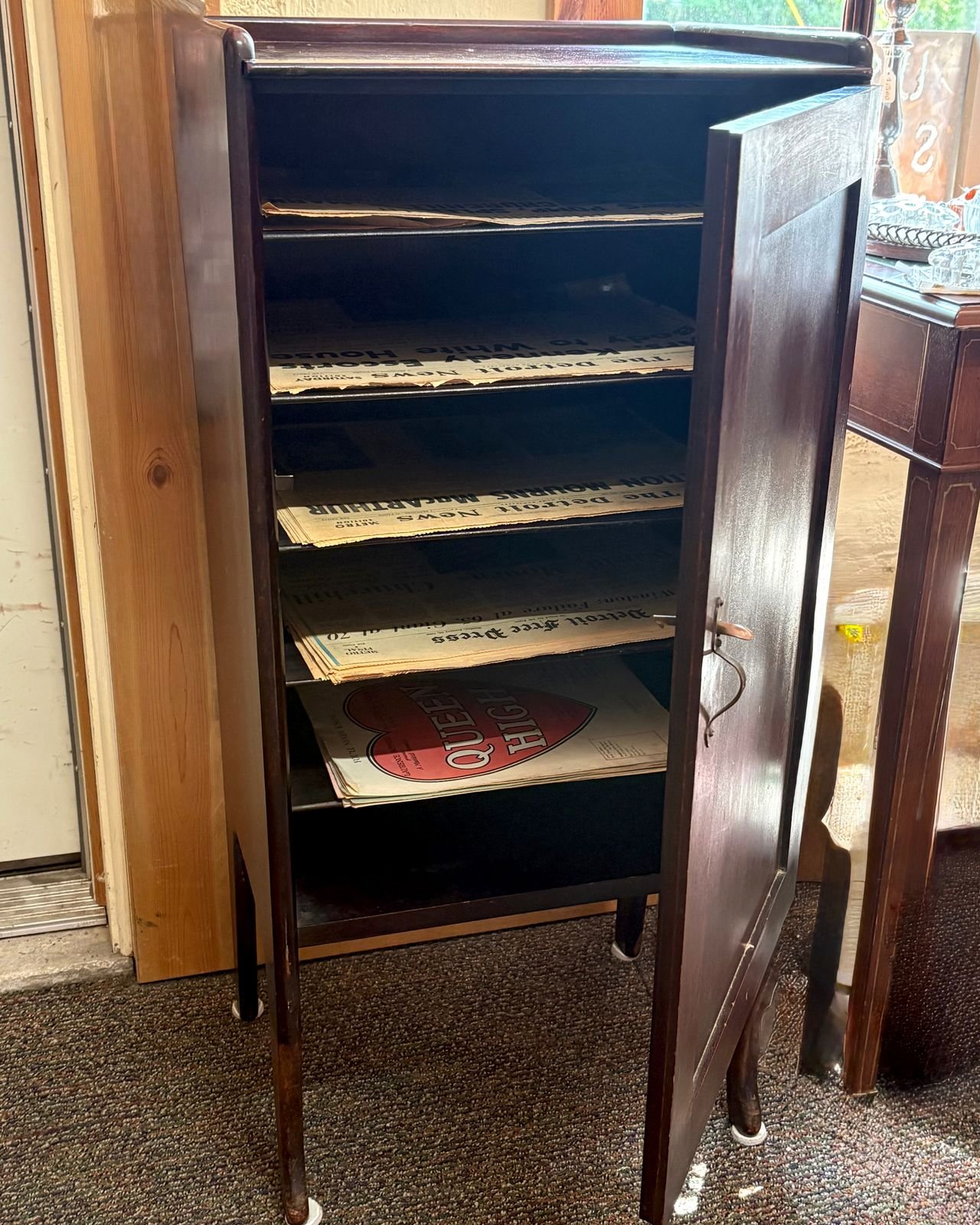 A tall narrow antique mahogany sheet music cabinet on four tapered legs, its single door standing open on fixed shelves stacked with vintage Detroit newspapers and sheet music.