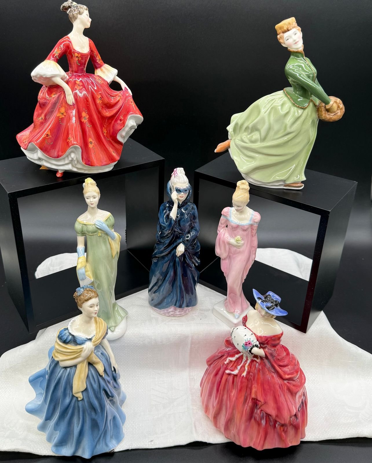 Seven Royal Doulton bone china lady figurines grouped on a black backdrop over a white cloth. A lady in a scarlet gown sprigged with orange flowers and a red-haired lady in a green jacket with a fur muff stand raised on black risers, and below them are ladies in sage green with long blue gloves, a dark blue masquerade cloak, pink holding a yellow rose, blue under a gold shawl, and a crimson crinoline with a wide blue hat.