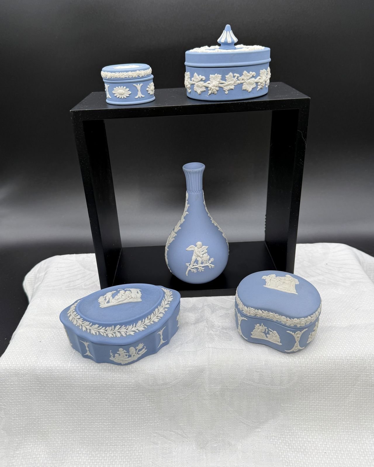 Five pieces of pale blue Wedgwood jasperware with white relief decoration, set on and around a black display box over a white cloth. On top sit a small drum-shaped lidded box banded with daisy rosettes and a round covered box wrapped in grapevine with a pointed white finial. A bud vase with a narrow fluted neck and a winged cupid relief stands inside the box frame, and in front are a scalloped oval lidded box with reclining figures inside a laurel wreath and a kidney-shaped lidded box ringed with a rose garland.