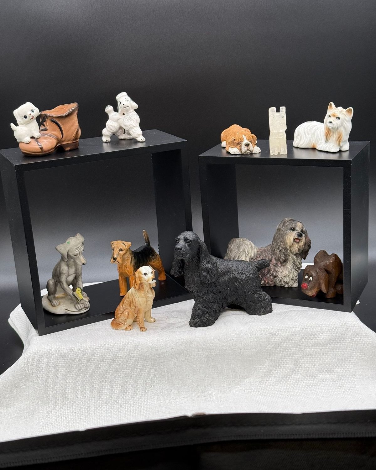 Eleven small dog figurines arranged on and around two black open display boxes set on a white cloth. On the left box sit a white puppy climbing into a brown ceramic boot and a standing white poodle, with a grey bisque dog on an oval base, a tan and black Airedale, and a seated golden retriever below. On the right box are a brown and white bulldog puppy lying down, a small pale carved stone dog, and a white and tan long haired terrier, with a black cocker spaniel, a shaggy grey terrier, and a dark carved wooden dog with a red nose in front.