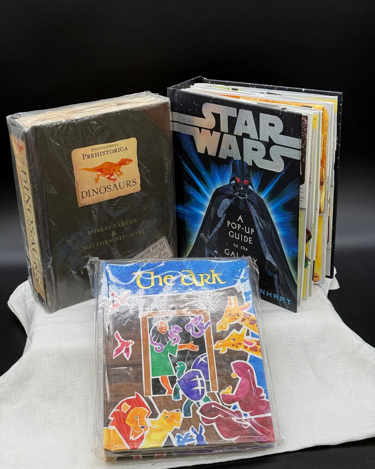 Three pop-up books arranged on a white cloth against a black backdrop. At left, Encyclopedia Prehistorica Dinosaurs by Robert Sabuda and Matthew Reinhart stands upright in a clear protective sleeve, its dark cover carrying a tan label with an orange tyrannosaurus and a sticker reading The Definitive Pop-Up. Behind it at right, Star Wars A Pop-Up Guide to the Galaxy stands open, showing Darth Vader on the cover and a thick fan of folded pages. In front sits The Ark, a pop-up by Matthew Reinhart, its blue and brown cover crowded with cut-paper lions, elephants, giraffes and birds.
