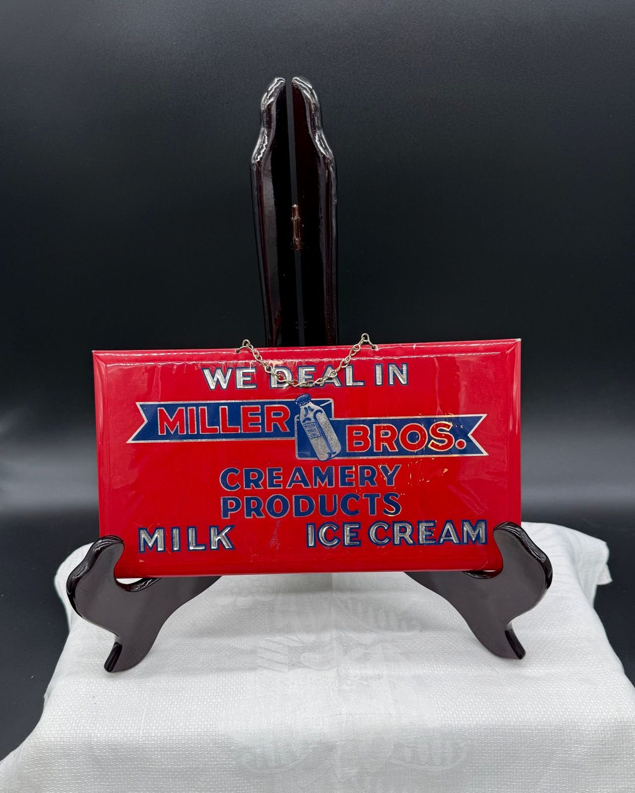A small rectangular embossed metal advertising sign with a bright red ground, resting on a dark wooden display easel on a white cloth against a dark grey backdrop. Silver embossed capitals at the top read WE DEAL IN. Below them a blue ribbon banner carries MILLER on the left and BROS. on the right, with a silver capped milk bottle standing between them. Beneath the banner, blue lettering reads CREAMERY PRODUCTS, and along the bottom MILK on the left and ICE CREAM on the right in silver. A short metal hanging chain crosses the top edge of the sign.