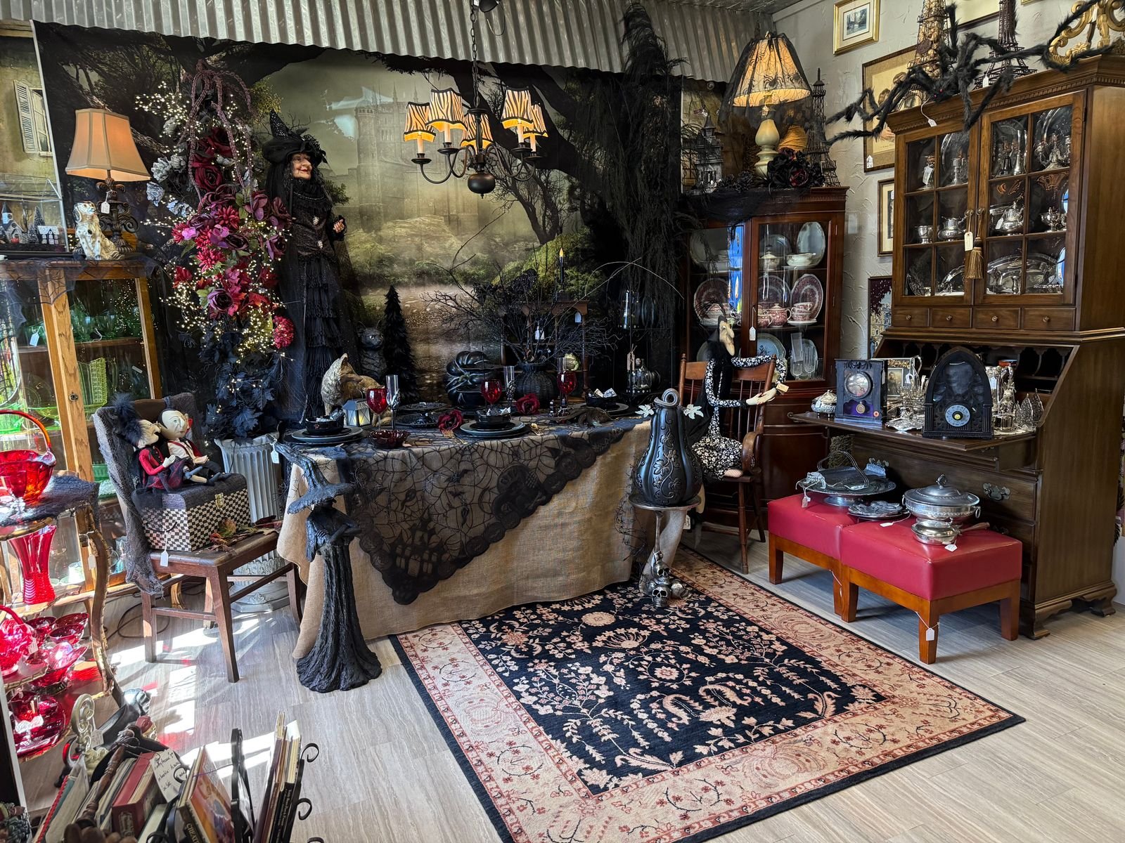 A Halloween dining display in an antique shop. A table draped in burlap and a black spiderweb lace cloth is set with black plates, ruby red goblets, and a coal-black pumpkin beneath a candle chandelier with striped shades. A tall witch in black lace and a pointed hat stands beside the table in front of a mural of a castle in a foggy wood. A second witch in a black and white dress sits at the head of the table. Two small vampire dolls share a wingback chair at left. Giant black spiders sit atop a wooden china cabinet full of silver at right, with a red leather bench holding silver serving pieces in front of it.