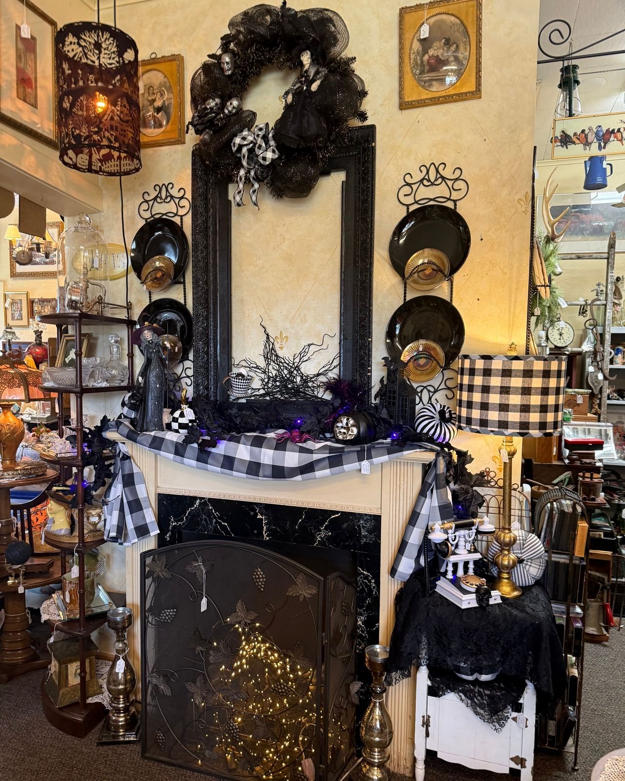 A Halloween fireplace mantel in an antique shop. A black mesh wreath studded with silver skulls and a pale figure in a black gown hangs over an empty carved black frame. Black plates backed by amber glass discs sit in scrolled wrought iron racks on either side. The mantel is draped in black and white buffalo check with black leaves, purple lights, a witch figurine in a violet hat, a skull pumpkin, a striped pumpkin, and a small pink spider. Below, a black marble surround holds a grapevine iron fireplace screen with tiny lights glowing behind it, flanked by tall silvered candlesticks. At right, a small white cabinet draped in black lace holds an ornate white rotary telephone with a black skull, beneath a buffalo check lamp.