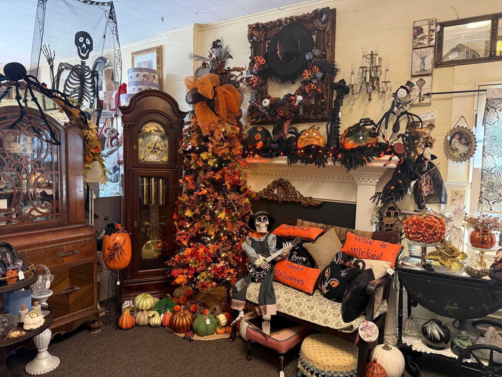 A large Halloween fireplace display in an antique shop. An ornate gilded frame on the wall holds a wreath with a wide black witch hat, orange polka-dot ribbons, and striped stockings dangling below. The white mantel is lined with black garland, orange lights, a grinning pumpkin face, a black glitter pumpkin, a spider holding a lollipop, and a top-hatted skeleton figure. Below, a skeleton in overalls plays a banjo on a settee piled with orange and black pillows reading Boo, Happy Halloween, and Wild and Wicked, plus a black cat pillow. At left stand a tall grandfather clock, an autumn tree with orange mesh bow and lights above a pile of pumpkins, and a waterfall-front cabinet with arched glass doors topped by a giant spider. A crystal candle sconce hangs on the wall at right beside a black drop-leaf tea cart.