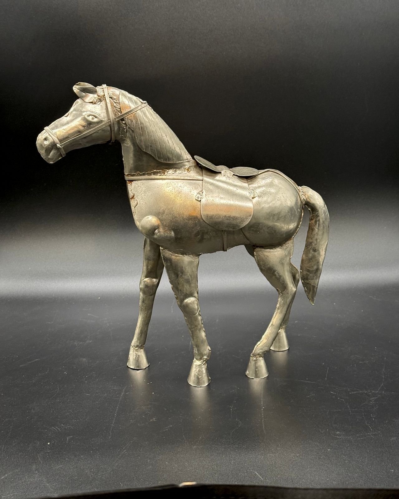 A handmade silvery sheet metal horse sculpture standing mid-stride against a dark backdrop. It has a wire bridle, a scored mane, a chest strap with small scroll ornaments, a saddle cloth with a small round saddle, domed knee caps, cone-shaped hooves, and visible soldered seams, with a warm patina showing hints of gold and copper.