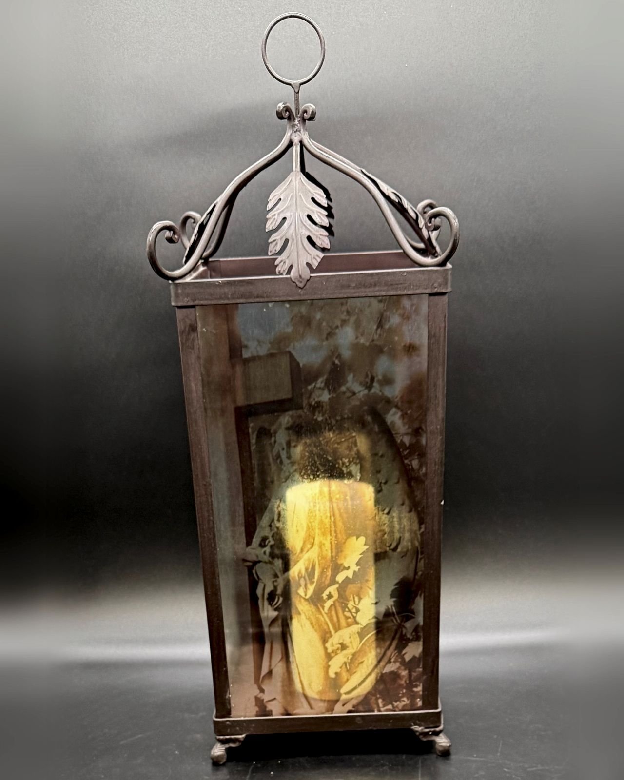 A tall metal and glass candle lantern against a dark gray backdrop. The dark bronze-toned frame has scrolled arms rising to a fleur-de-lis finial and round hanging ring, a metal leaf ornament at the top center, and four small scrolled feet. The glass panel shows a sepia and softly tinted image of a stone angel beside a cross among autumn leaves, glowing gold where a pillar candle is lit inside.