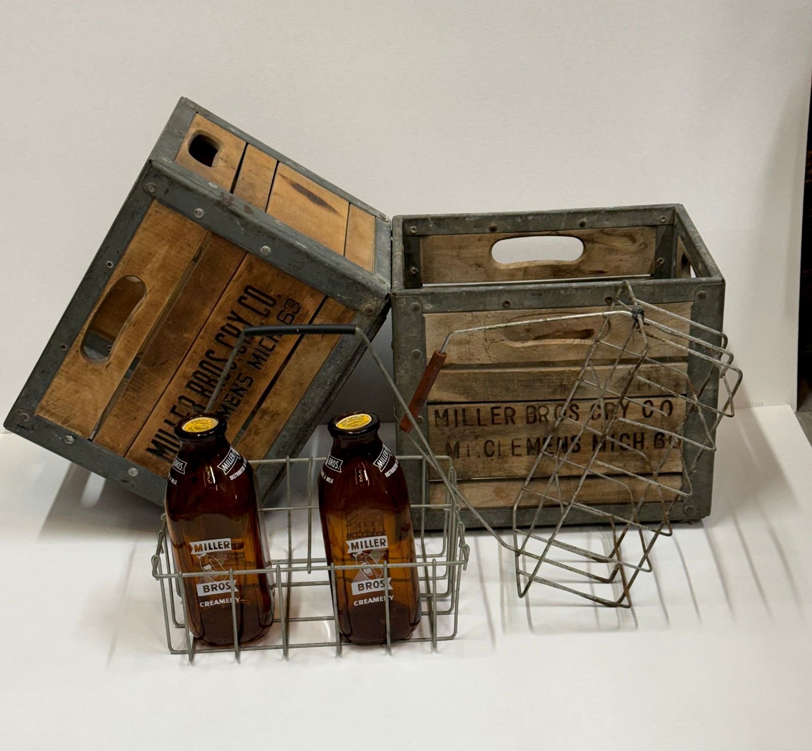 Two weathered wooden milk crates with galvanized steel trim stenciled Miller Bros Cry Co Mt. Clemens Mich., two amber Miller Bros Creamery bottles in a wire carrier, and a second wire bottle carrier with a red wooden handle.