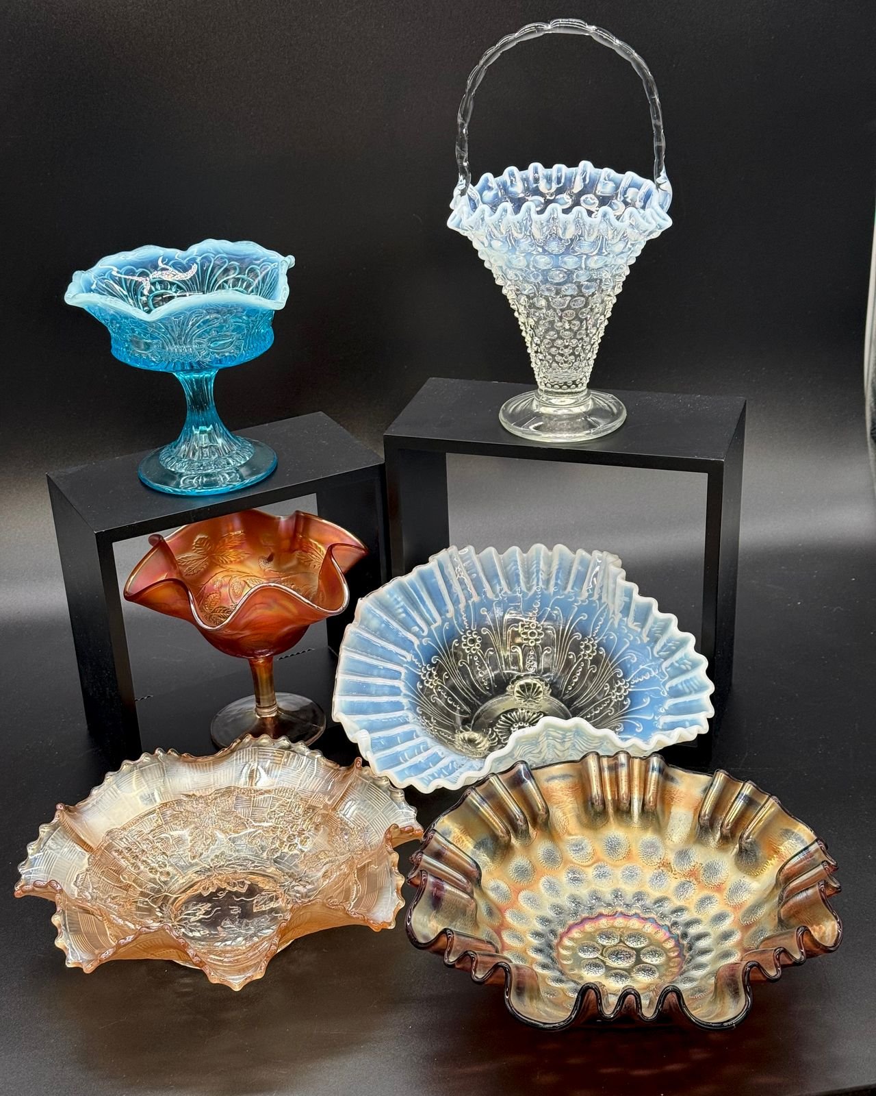 Six pieces of vintage glass on black risers: a blue opalescent compote, an opalescent hobnail basket with a clear handle, a marigold carnival compote, a ruffled opalescent bowl, a peach carnival bowl, and an iridescent purple-edged carnival bowl.