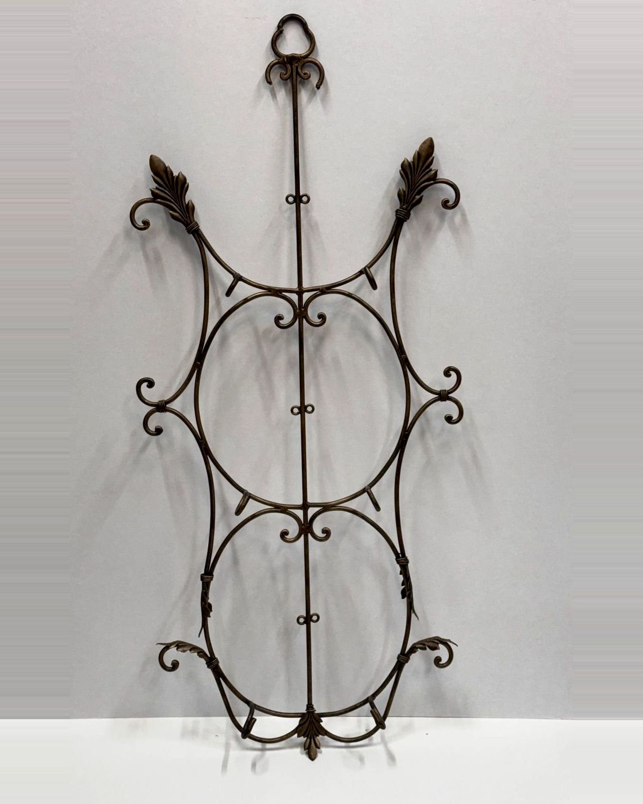 Tall vintage brass wall plate rack with a hanging ring, leaf finials and scrollwork, with hooks and clips to hold three large plates stacked vertically.