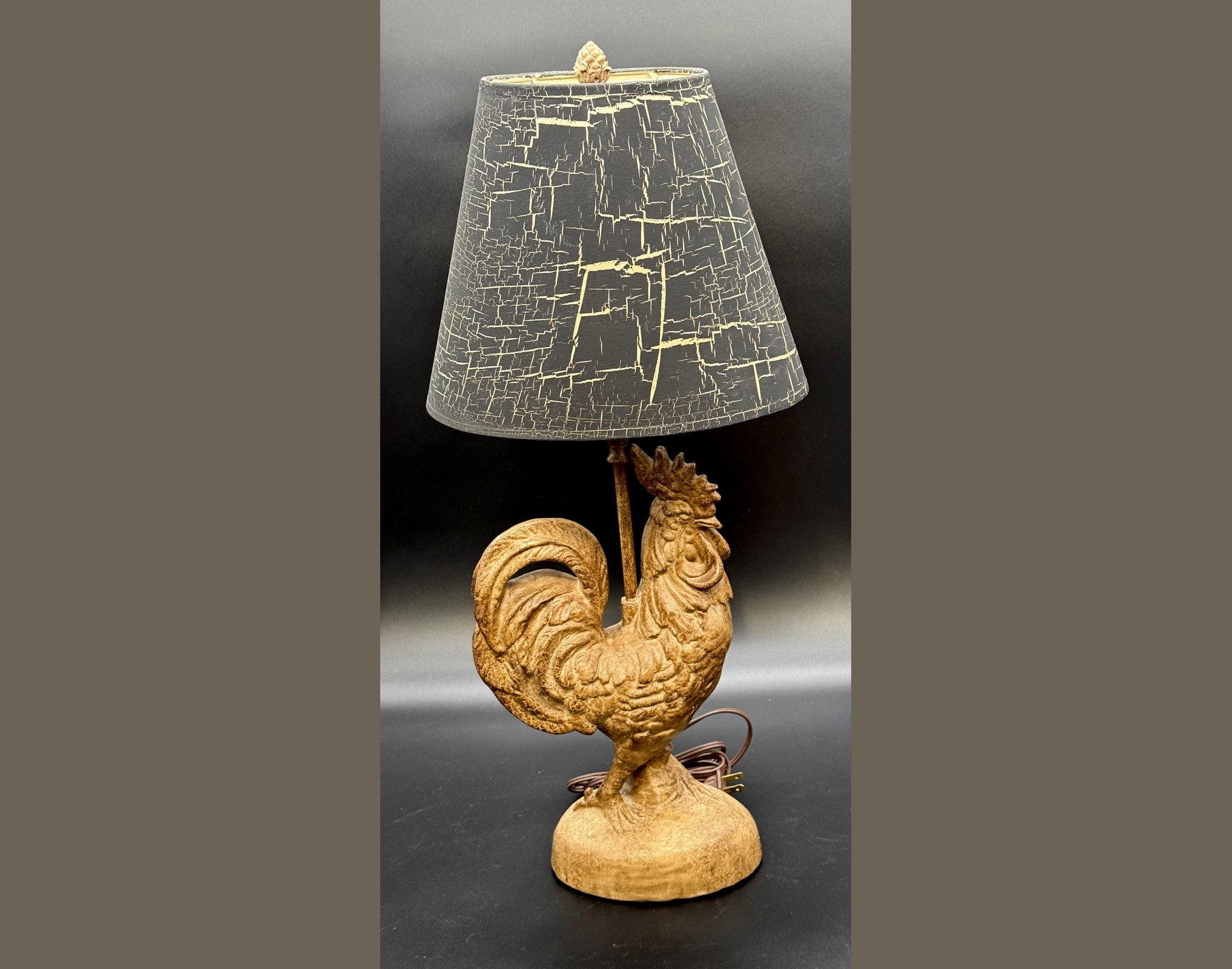 A vintage sculpted rooster table lamp with a sun-warmed aged finish on a cast rooster figure standing on a round base, topped with a tapered black drum lampshade featuring a striking ivory crackle texture pattern.
