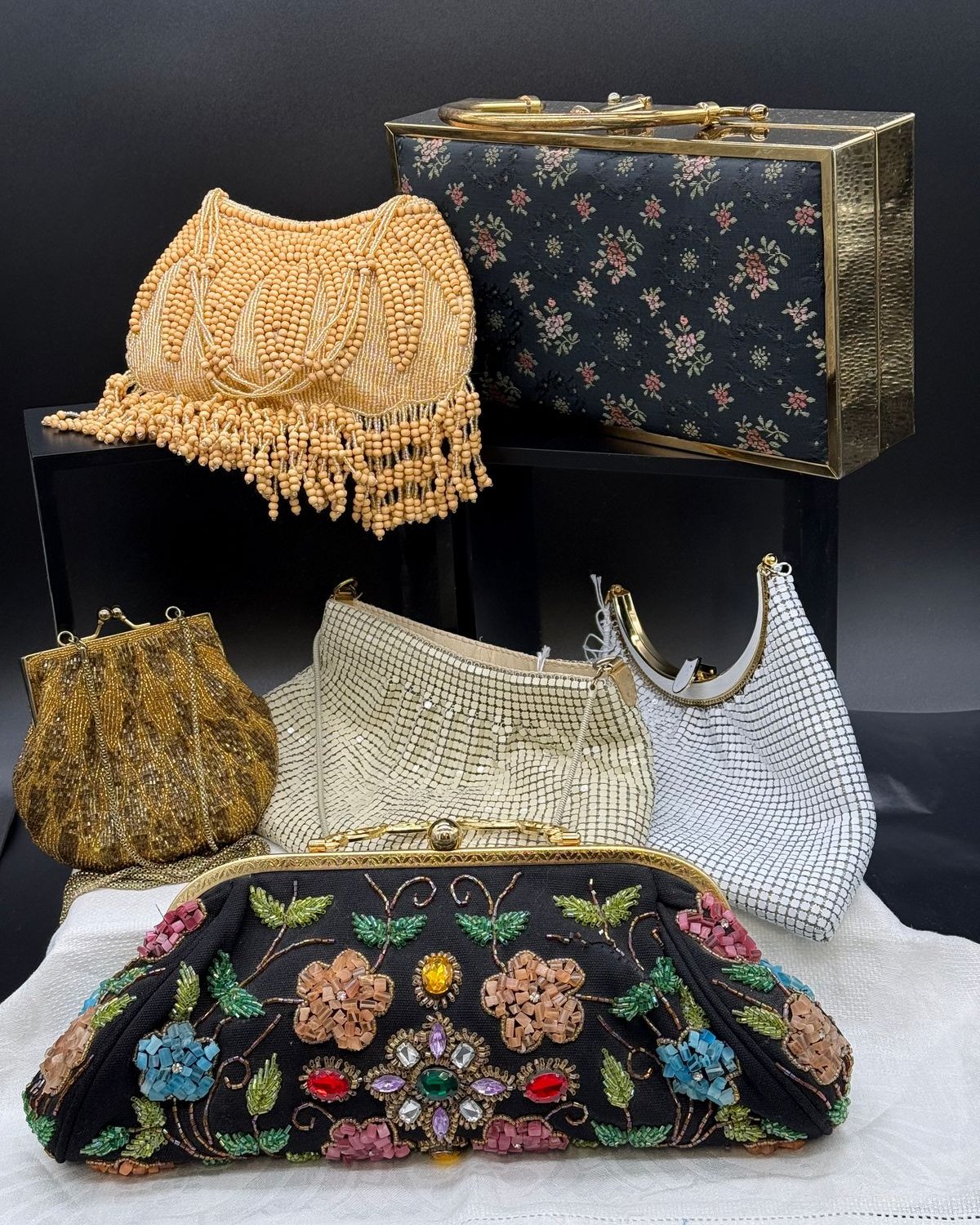 A collection of six vintage purses arranged together: a black evening clutch covered in jeweled multicolor floral beadwork, a cream and peach beaded evening bag with long bead fringe, a small gold metallic beaded coin purse, an ivory chainmail mesh bag, a silver chainmail mesh bag with gold frame, and a black floral box purse trimmed in gold.